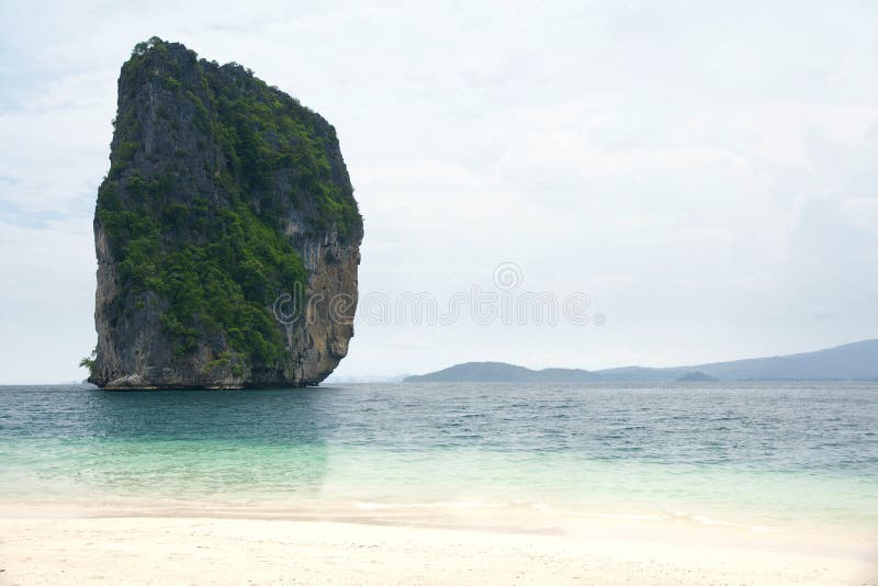 Big high rock cliff filled with green vegetation surrounded by turquoise blue colored ocean water next to a tropical white sand beach with horizon view at midday, Krabi Thailand. Big high rock cliff filled with green vegetation surrounded by turquoise blue colored ocean water next to a tropical white sand beach with horizon view at midday, Krabi Thailand.