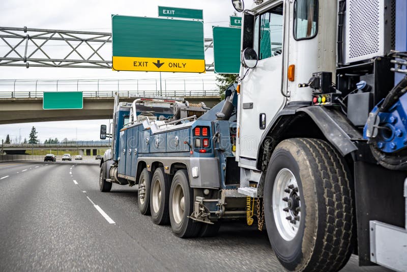 Big rig powerful tow semi truck towing broken truck on the lifting boom driving on the wide highway road with road signs and