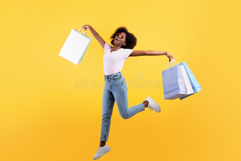Big sales concept. Young african american lady jumping with colorful shopping bags over yellow background