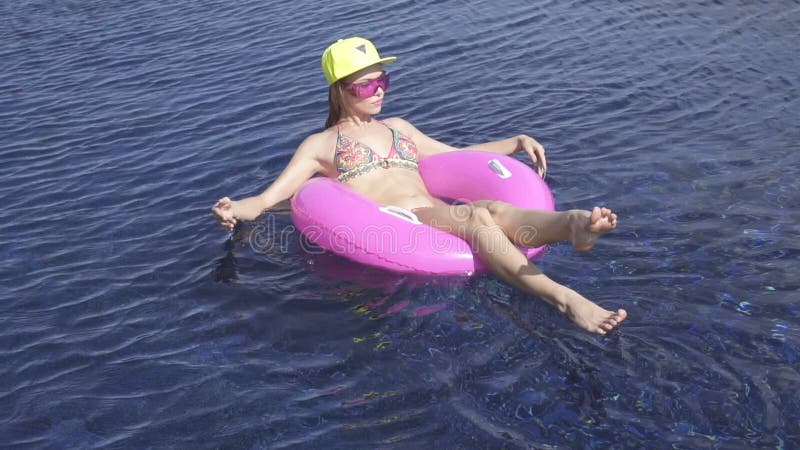 Bikini d'uso della giovane donna graziosa, cappello giallo ed occhiali da sole porpora sedentesi su un anello gonfiabile rosa nel