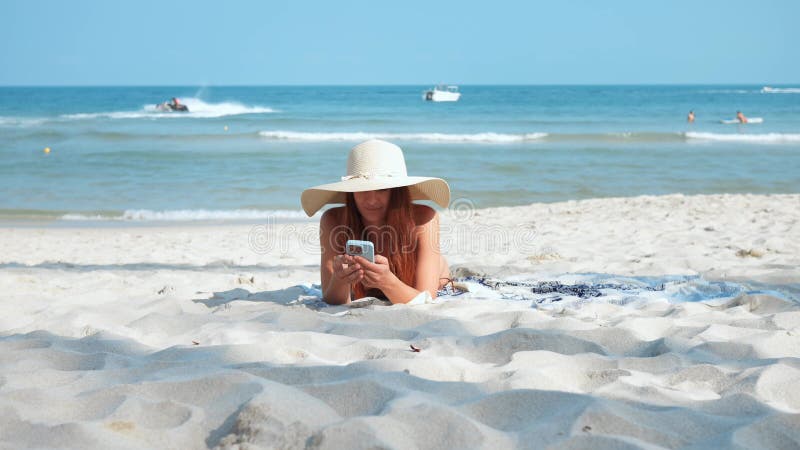 In bikini and sun hat woman lying on sandy beach using phone vacation. Vacation on beach sun's gentle warmth finds