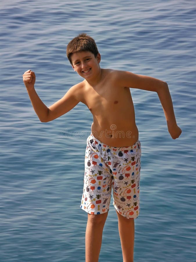 Smiling boy in swim wear shows her strength ( muscles) near the blue Adriatic sea at sunny day. Vertical color photo. Smiling boy in swim wear shows her strength ( muscles) near the blue Adriatic sea at sunny day. Vertical color photo.