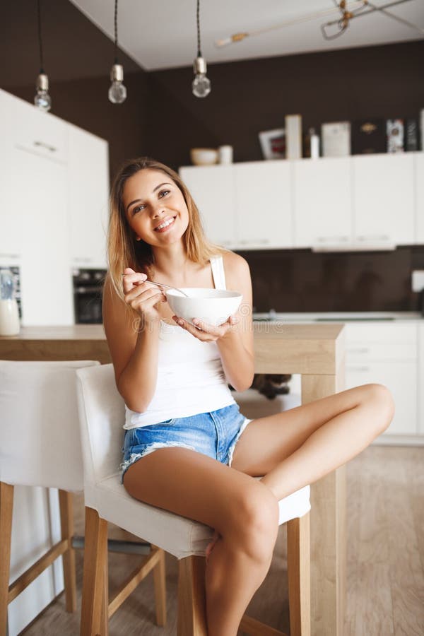 Bilden av den attraktiva unga flickan som äter cornflakes med, mjölkar på kök som ser att le för kamera royaltyfria bilder Bilden av den attraktiva unga flickan som äter cornflakes med, mjölkar på kök som ser att le för kamera royaltyfria bilder