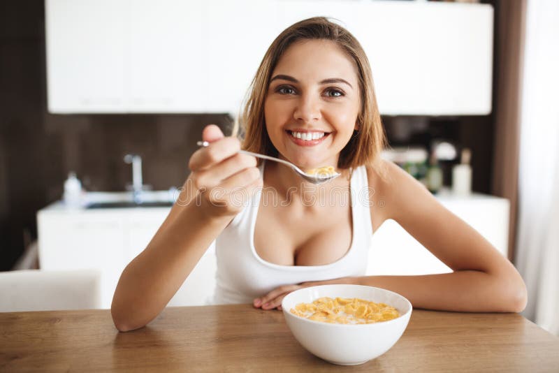 Bilden av den attraktiva unga flickan som äter cornflakes med, mjölkar på köklookig på att le för kamera arkivfoton Bilden av den attraktiva unga flickan som äter cornflakes med, mjölkar på köklookig på att le för kamera arkivfoton