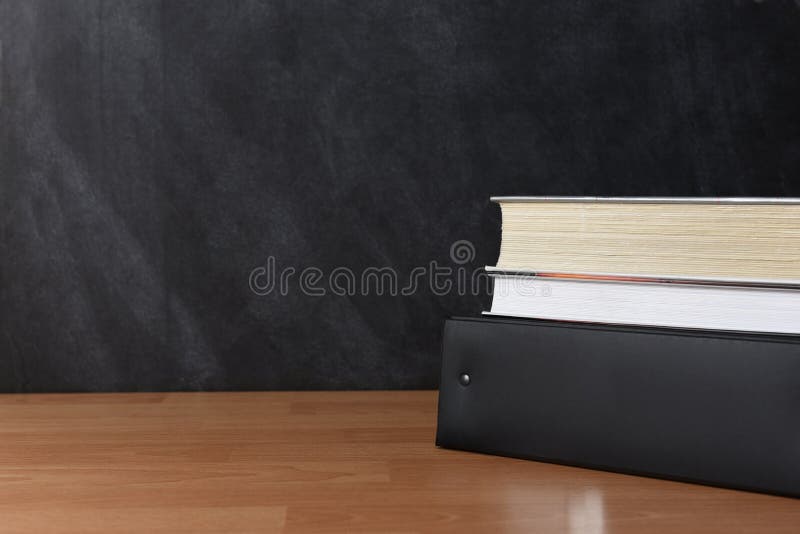 A binder with two textbooks on a teachers desk in front of a chalkboard