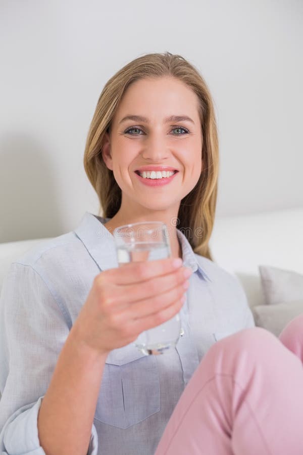 Bionda casuale felice che si siede sullo strato che tiene bicchiere d'acqua immagine stock Bionda casuale felice che si siede sullo strato che tiene bicchiere d'acqua immagine stock
