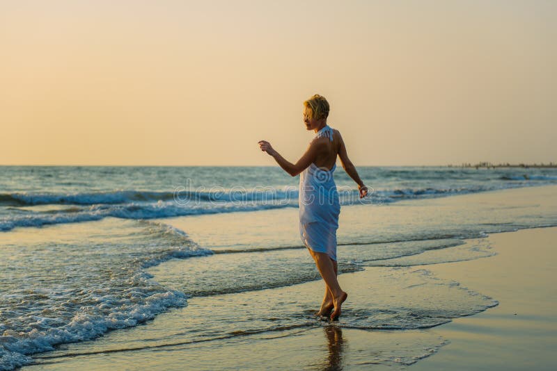Bionda snella incantante nei supporti alla moda del vestito in onde sul mare La giovane donna cammina a piedi nudi lungo la spuma immagini stock Bionda snella incantante nei supporti alla moda del vestito in onde sul mare La giovane donna cammina a piedi nudi lungo la spuma immagini stock