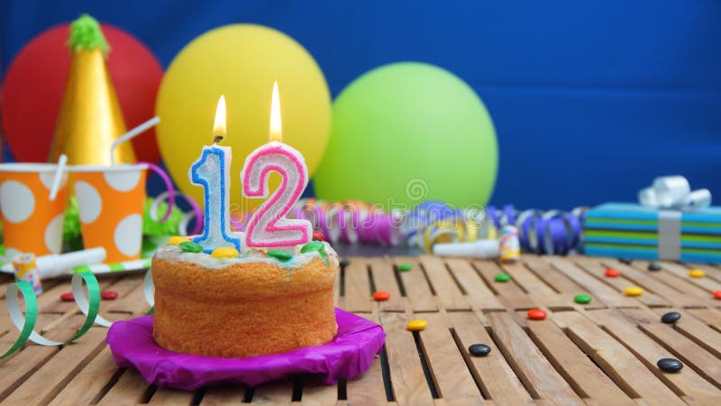 Birthday 12 cake with candles on rustic wooden table with background of colorful balloons, gifts, plastic cups and candies