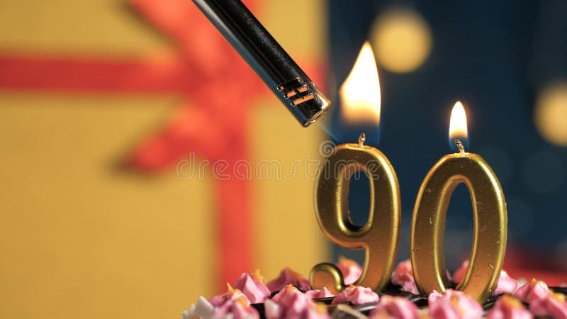 Birthday cake number 90 golden candles burning by lighter, background gift yellow box tied up with red ribbon. Close-up