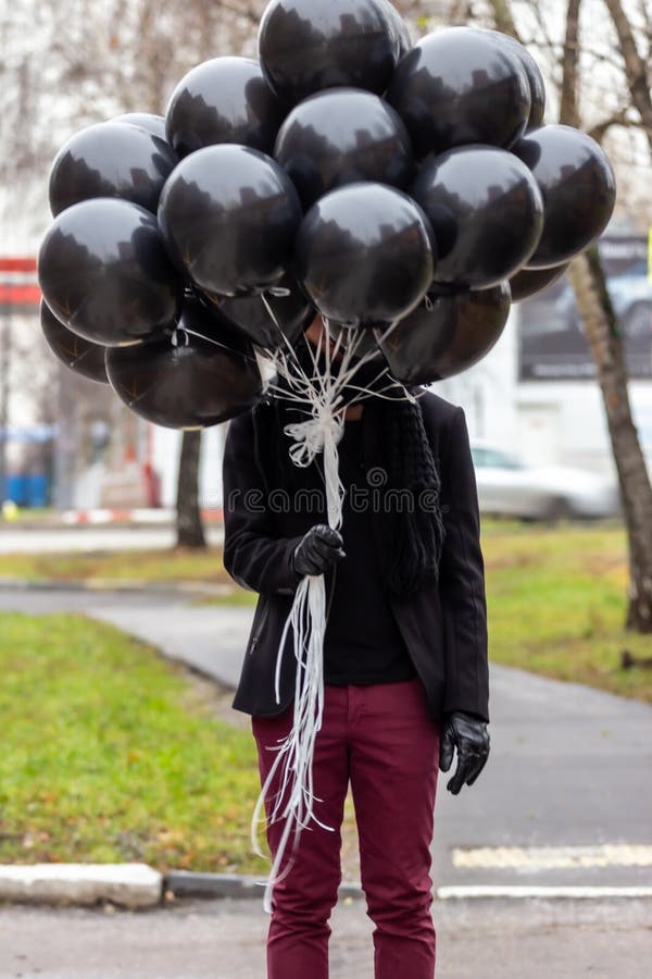 Birthday is a sad holiday concept. A young man wearing black scarf and coat hiding behind black balloons, blur and grain effect.