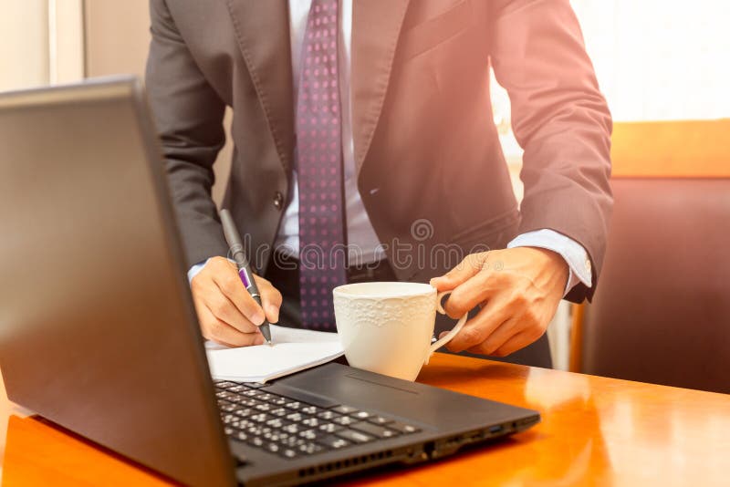 Biznesmen ręka trzyma filiżankę i pisze na notatniku na wprking biurku zdjęcie royalty free Biznesmen ręka trzyma filiżankę i pisze na notatniku na wprking biurku zdjęcie royalty free