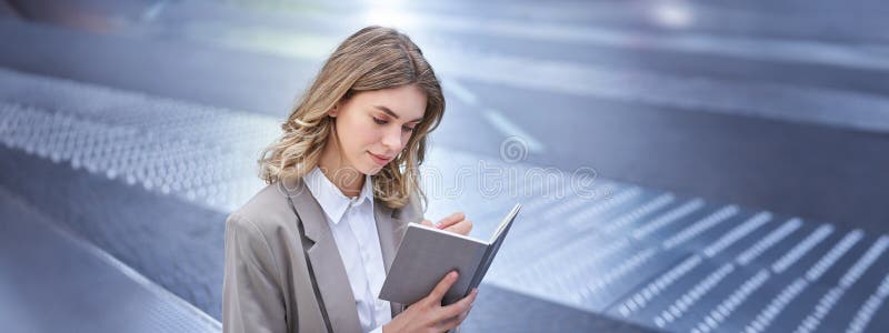 Businesswoman in corporate suit, writes down smth, works on ideas in notebook, sits outside in city centre. Businesswoman in corporate suit, writes down smth, works on ideas in notebook, sits outside in city centre.