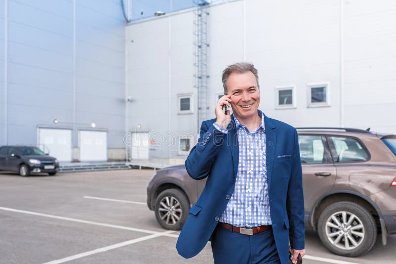 Biznesmen w średnim wieku w niebieskim garniturze śmiejący się podczas rozmowy telefonicznej wychodzi z samochodu na prywatny par zdjęcie stock Biznesmen w średnim wieku w niebieskim garniturze śmiejący się podczas rozmowy telefonicznej wychodzi z samochodu na prywatny par zdjęcie stock