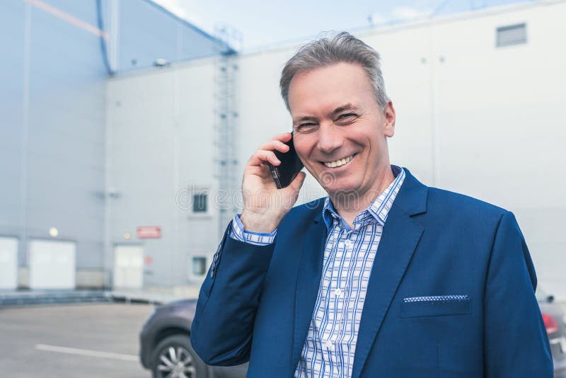 Biznesmen w średnim wieku w niebieskim garniturze śmiejący się podczas rozmowy telefonicznej wychodzi z samochodu na prywatny par zdjęcie stock Biznesmen w średnim wieku w niebieskim garniturze śmiejący się podczas rozmowy telefonicznej wychodzi z samochodu na prywatny par zdjęcie stock