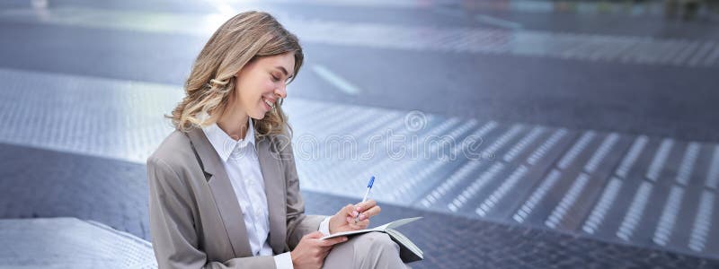 Business woman in suit sits in city centre, writes down, takes notes, holds pen and notebook, brainstorms, creates ideas. Business woman in suit sits in city centre, writes down, takes notes, holds pen and notebook, brainstorms, creates ideas.