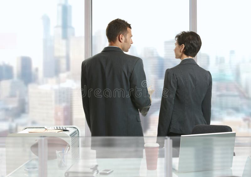 Two businesspeople standing at desk in office, looking out the windows and talking. Two businesspeople standing at desk in office, looking out the windows and talking.