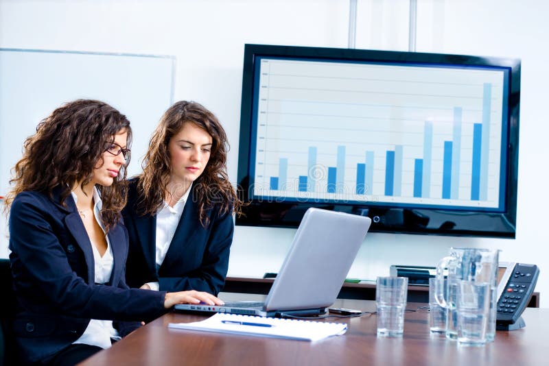 Young businesswomen sitting by meeting table at office in front of a huge blank plasma TV screen and using laptop and phone. Young businesswomen sitting by meeting table at office in front of a huge blank plasma TV screen and using laptop and phone.