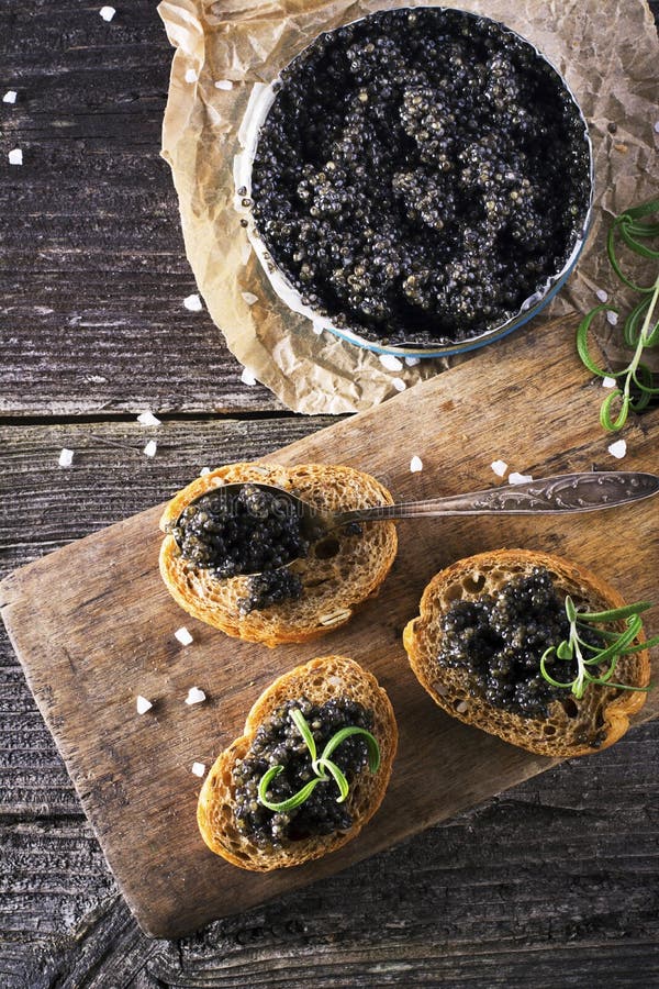 Black Astrakhan Russian salted sturgeon caviar in a can on dark background with wooden spoon and slices of bread. Snacks