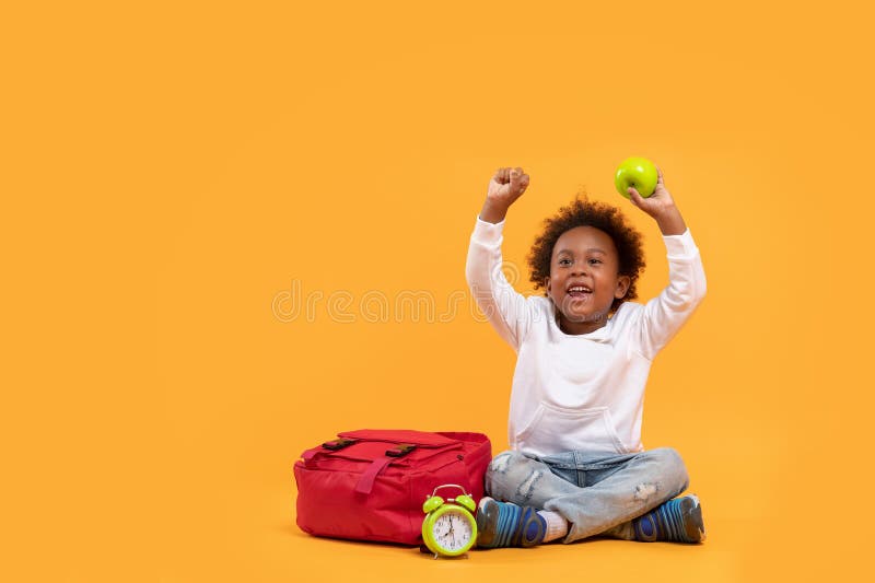 Black child boy 3 years, Student kid holding green apple in his hand and Yay! happy while sitting with school bag and alarm clock