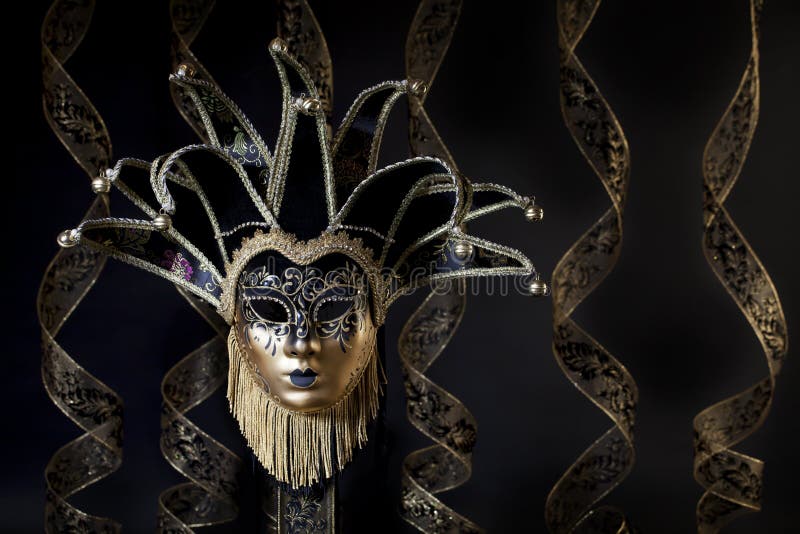 A black and gold Venetian Jester style mask on black background with gold and black curled ribbon streamers blurred in background. A black and gold Venetian Jester style mask on black background with gold and black curled ribbon streamers blurred in background