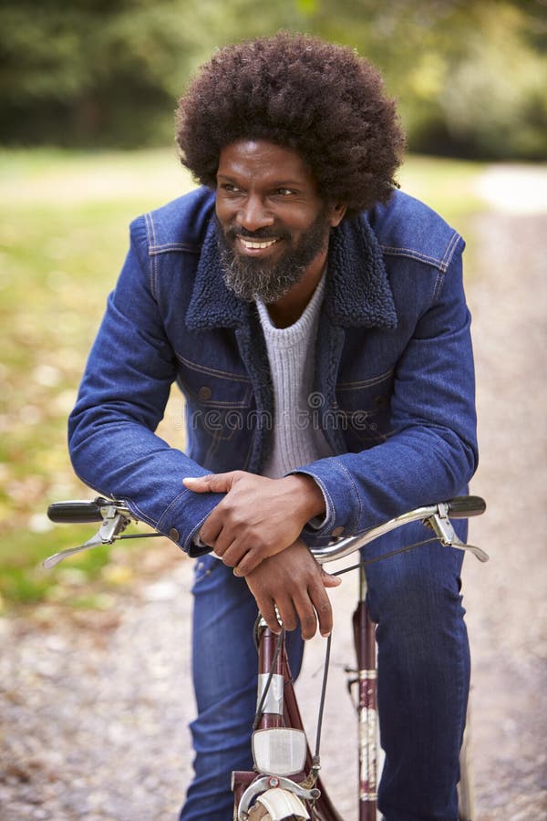 Black middle aged man sitting on a bike in a park, leaning on the handlebars smiling, front view, close up
