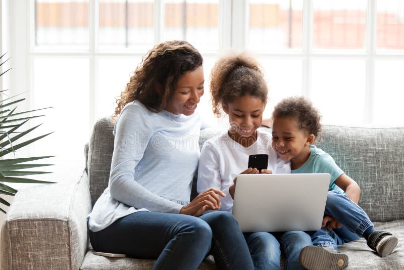 Young black family sit on couch at home using devices playing game on smartphone, millennial mom have fun spending time with mixed race kids hold laptop and cellphone. Children and technology concept. Young black family sit on couch at home using devices playing game on smartphone, millennial mom have fun spending time with mixed race kids hold laptop and cellphone. Children and technology concept