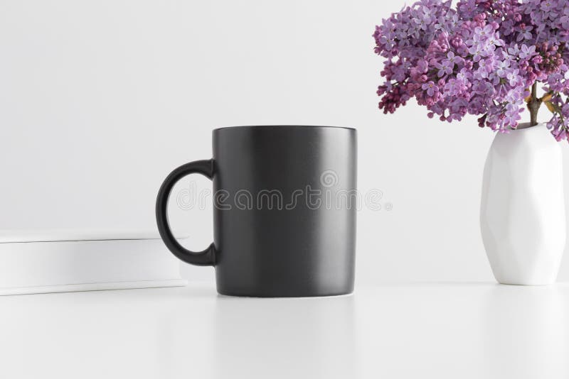 Black mug mockup with a book and a bouquet of lilac in a vase on a white table