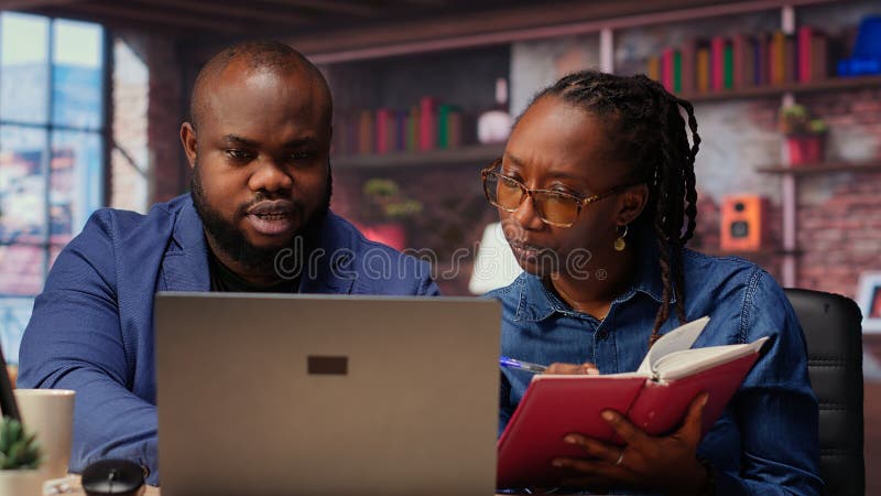 Black remote employees organizing calendar appointments using laptop and ledger stock photo