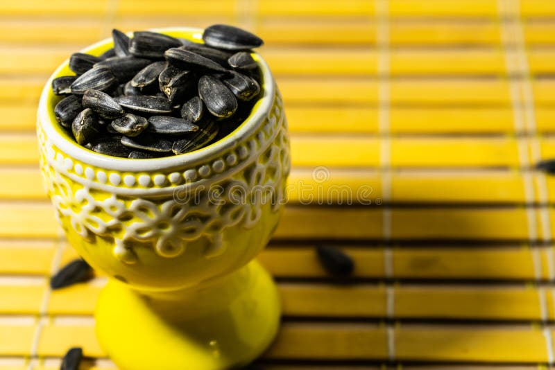 Black small sunflower seeds. Click seeds with husks. A handful in a yellow miniature stand on a wooden napkin. Spilled some seeds. Round small bowl with an openwork pattern.