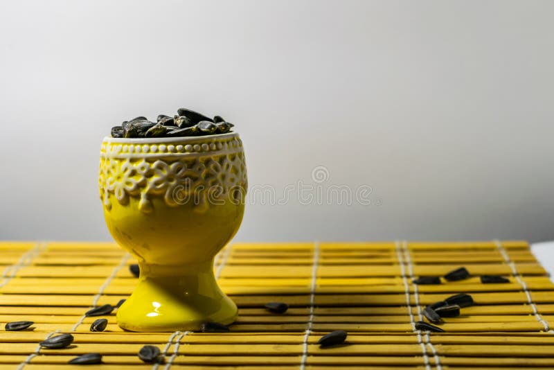 Black small sunflower seeds. Click seeds with husks. A handful in a yellow miniature stand on a wooden napkin. Spilled some seeds. Round small bowl with an openwork pattern.