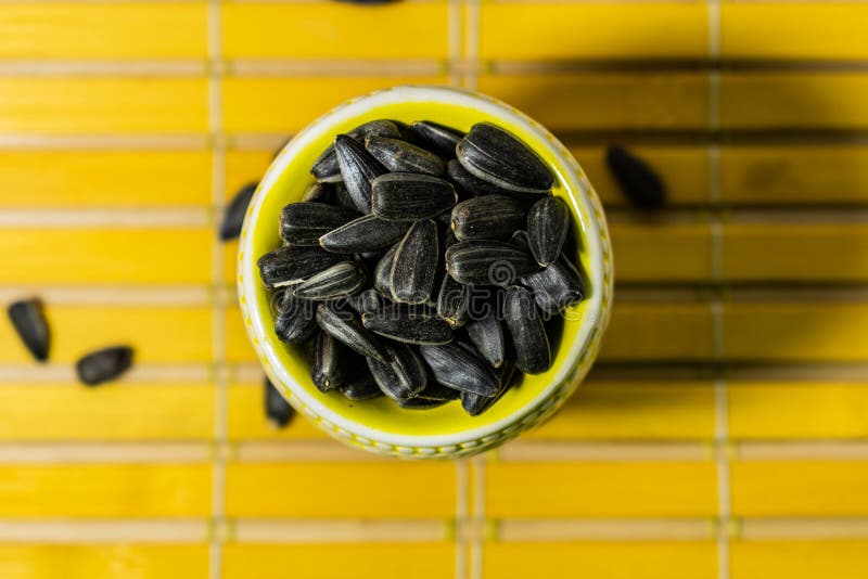 Black small sunflower seeds. Click seeds with husks. A handful in a yellow miniature stand on a wooden napkin. Spilled some seeds. Round small bowl with an openwork pattern.