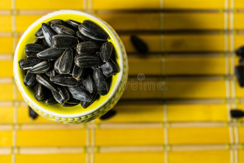 Black small sunflower seeds. Click seeds with husks. A handful in a yellow miniature stand on a wooden napkin. Spilled some seeds. Round small bowl with an openwork pattern.