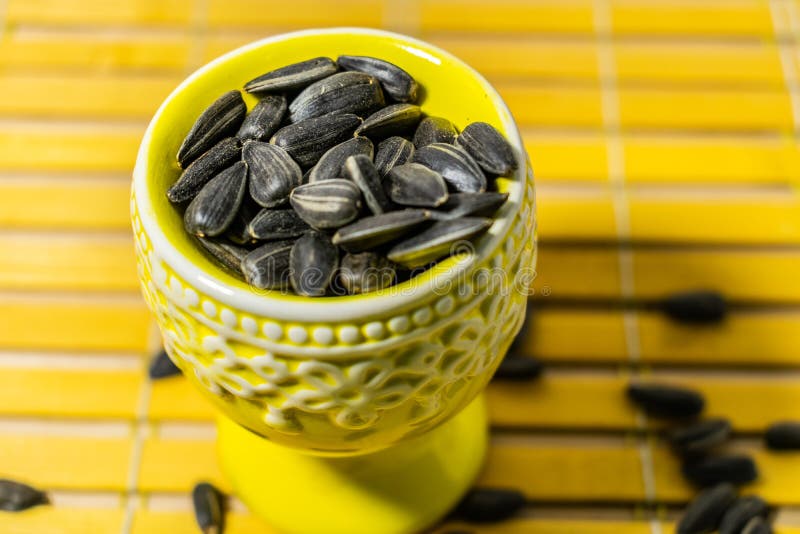 Black small sunflower seeds. Click seeds with husks. A handful in a yellow miniature stand on a wooden napkin. Spilled some seeds. Round small bowl with an openwork pattern.