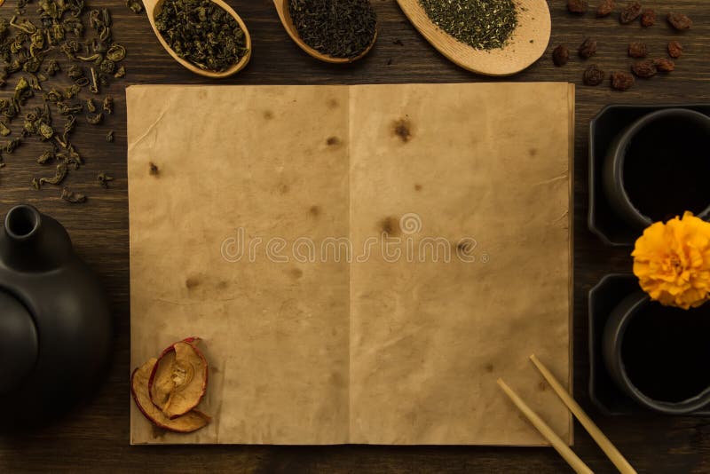 Black teapot, two cups, a collection of tea, dried apples, old blank open book on wooden background.