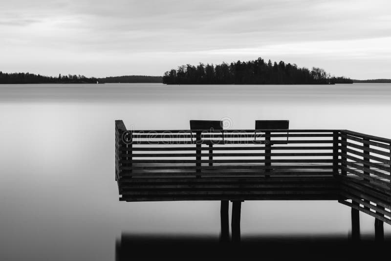 Black and white tranquil scenery of a lake with pier and two chairs. Water surface looks foggy due to long exposure.