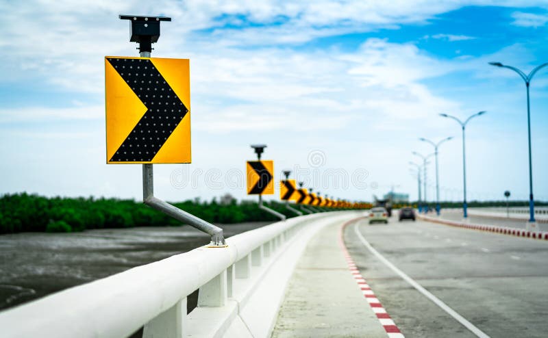 Black and yellow arrow on curve traffic sign on the bridge with solar cell panel ob blurred background of concrete road and car