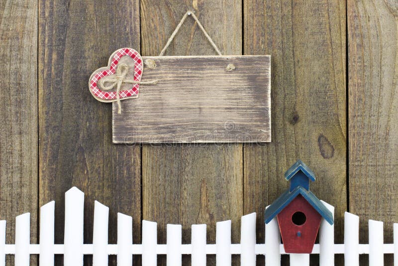 Blank wood sign with plaid heart hanging over white picket fence with birdhouse