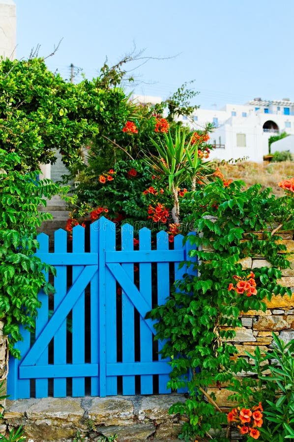 Painted blue garden gate surrounded by flowers and greenery with Mediterranean white washed buildings in background, Cyclades, Greece. Painted blue garden gate surrounded by flowers and greenery with Mediterranean white washed buildings in background, Cyclades, Greece.