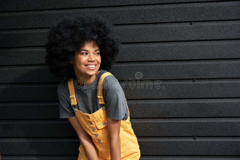 Happy stylish African American young woman wearing trendy sundress with afro hair looking away standing against black wall. Smiling mixed race generation z hipster lady posing on outdoor background. Happy stylish African American young woman wearing trendy sundress with afro hair looking away standing against black wall. Smiling mixed race generation z hipster lady posing on outdoor background.