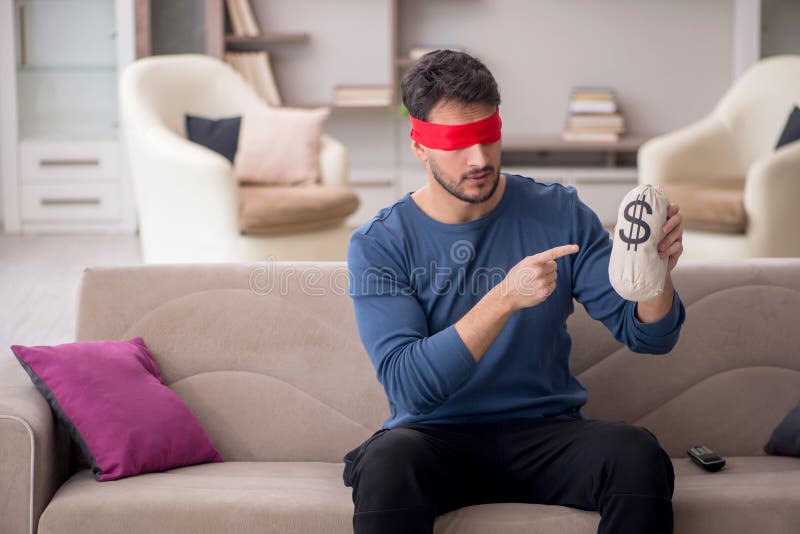 Young blindfolded man holding moneybag at home