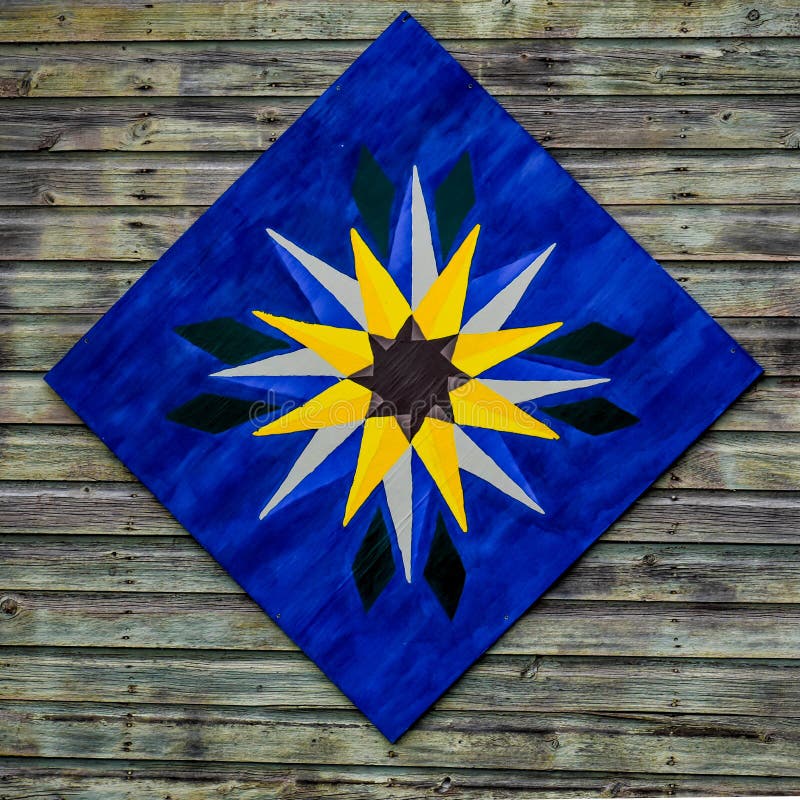 A painted quilt block hanging on a wooden barn.  The quilt pattern is blue with a star shaped pattern of golden yellow. A painted quilt block hanging on a wooden barn.  The quilt pattern is blue with a star shaped pattern of golden yellow.