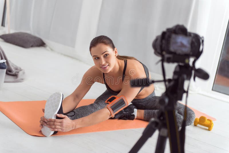 Being healthy. Attractive alert muscular dark-haired young blogger smiling and stretching while sitting on the carpet and making a video for her blog. Being healthy. Attractive alert muscular dark-haired young blogger smiling and stretching while sitting on the carpet and making a video for her blog