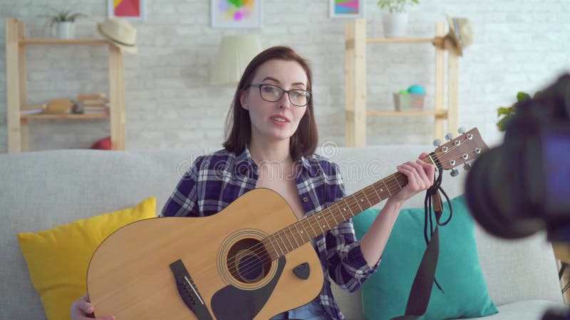 Blogger de jeune femme de portrait s'asseyant sur le divan avec une guitare dans des ses mains devant la caméra banque de vidéos