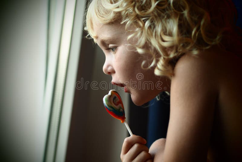 Blond błękit przyglądająca się dziewczynka z cukierkiem out w jej ręce przyglądającej okno zdjęcie stock Blond błękit przyglądająca się dziewczynka z cukierkiem out w jej ręce przyglądającej okno zdjęcie stock
