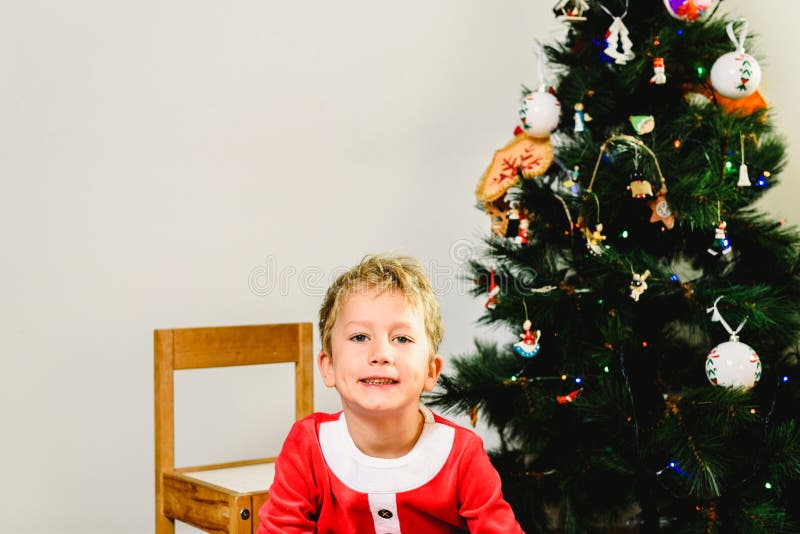 Blond and handsome 5 year old boy dressed up as santa claus next to christmas tree and white background with space for text