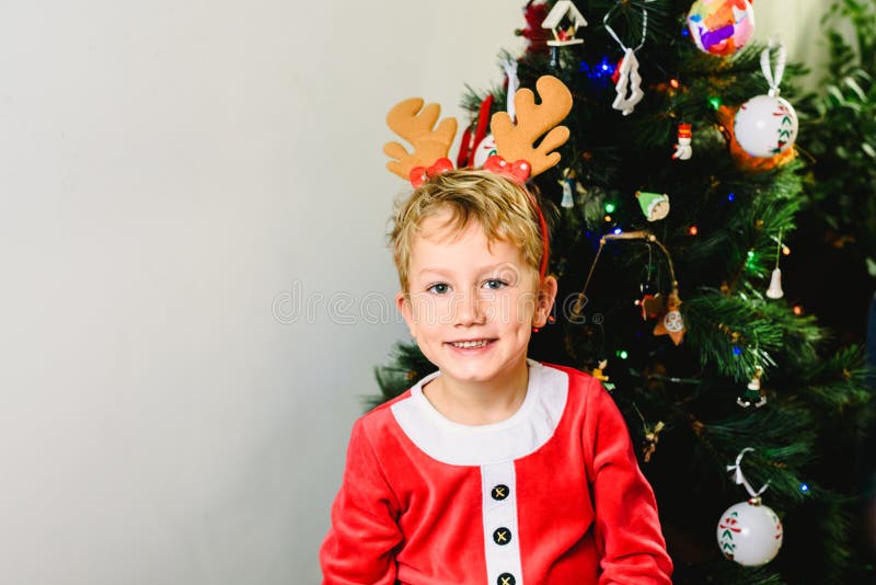 Blond and handsome 5 year old boy dressed up as santa claus next to christmas tree and white background with space for text