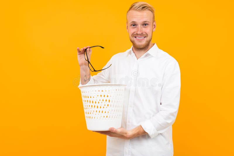 Blond man dressed in a classic white shirt throws glasses in the trash bin on a yellow background