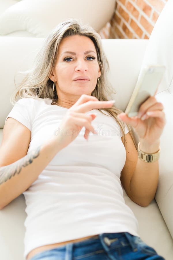Blond woman with tattoo in jeans using smart phone lying on white sofa stock photos