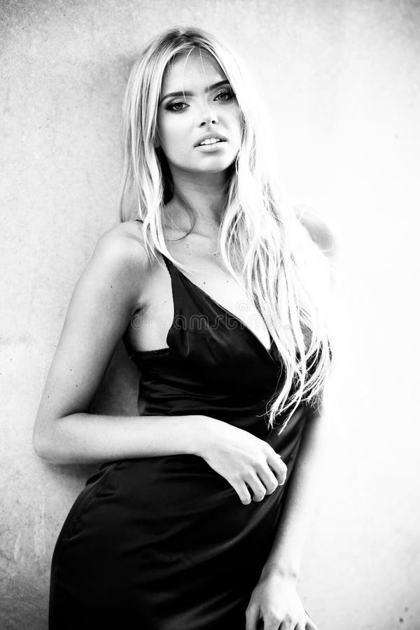 Blond young woman with beautiful makeup and hair in black dress standing near grey wall. Fashion model posing in elegant