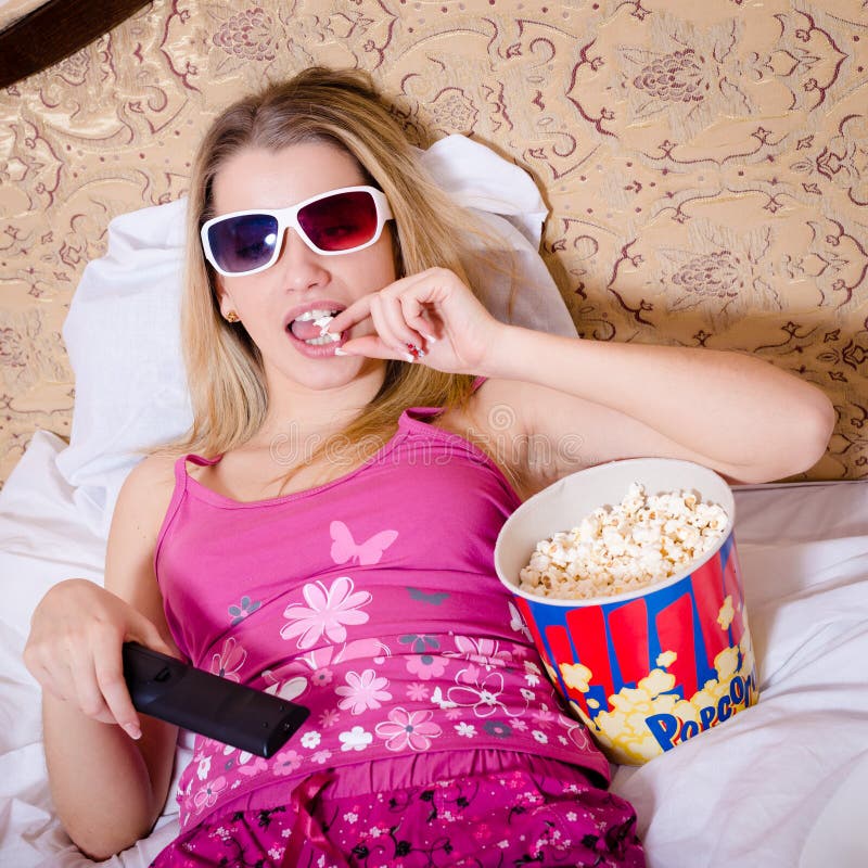 Blond young woman in color pajamas lying in bed with TV remote control in hand watching movie in 3D glasses and eating popcorn